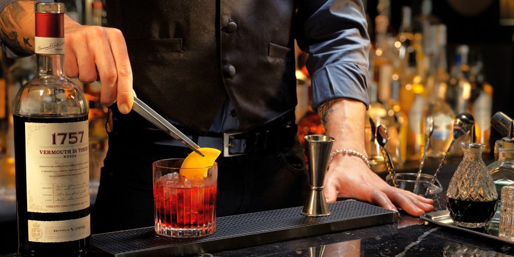 A bartender garnish a Negroni cocktail with a wedge of orange with a bottle of 1757 Vermouth di Toroni Rosso in the foreground