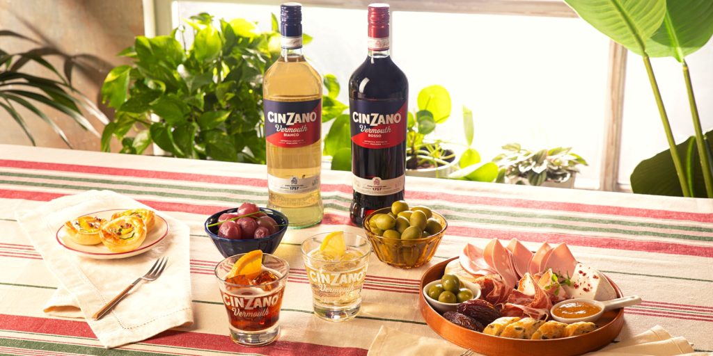 A bottle of Cinzano Vermouth Bianco and Cinzano Vermouth Rosso on a table with snacks and cocktails