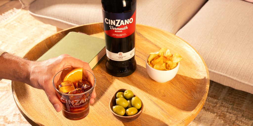 A bottle of Cinzano Vermouth Rosso on a table with a bowl of olives and a bowl of crisps
