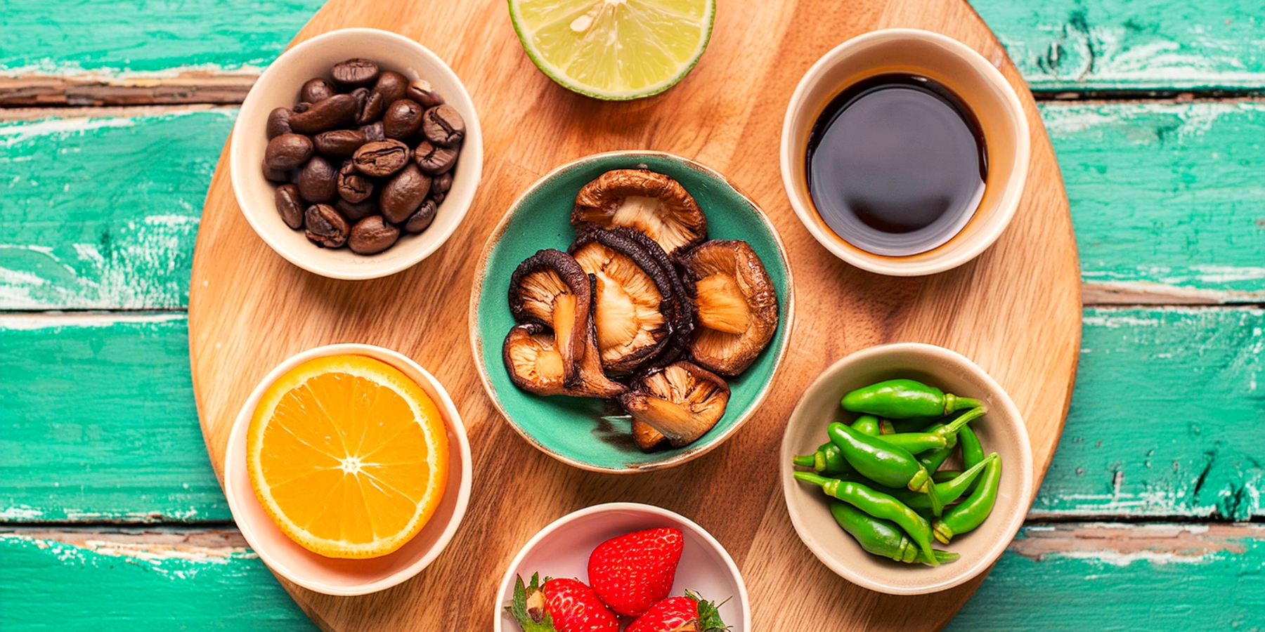 An overhead shot of small bowls of ingredients including soy sauce, coffee beans, green chillies, strawberries, fresh lime and fresh orange, arranged in a circle on top of a wooden board