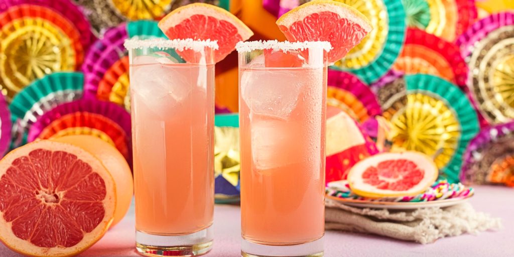 Two tall and refreshing light pink Paloma cocktails with a Cinco de Mayo theme in the background