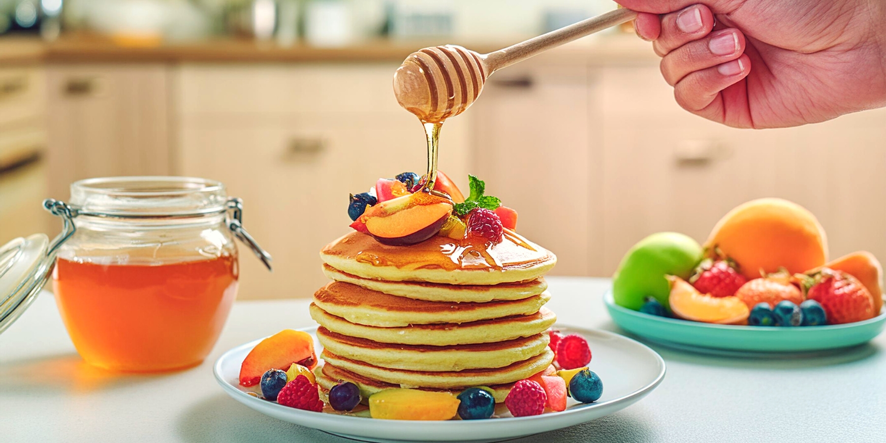 A hand holding a honey spoon and drizzling honey over a stack of pancakes and fruit