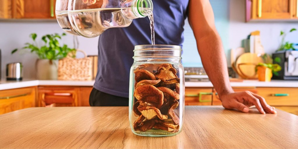 Close up view of a man pouring vodka over dried shitake mushrooms in a mason jar, in a light, bright modern kitchen