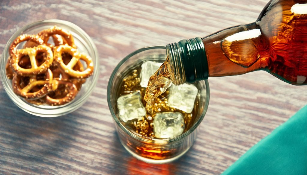 A top view of bourbon being poured into an ice-filled glass, bowl of pretzels to the side