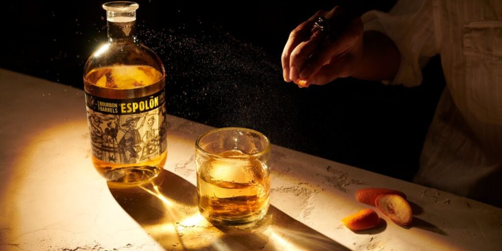 Espolòn Old Fashioned on a wooden table, with a hand squeezing a lemon peel, releasing its oils into the cocktail