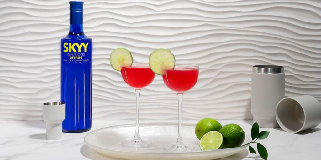 A vibrant cosmopolitan cocktail with lime garnishes sits next to a SKYY Vodka bottle, set against a white textured background. The red cocktail pops against the clean, modern setting, embodying a chic, feminine vibe.
