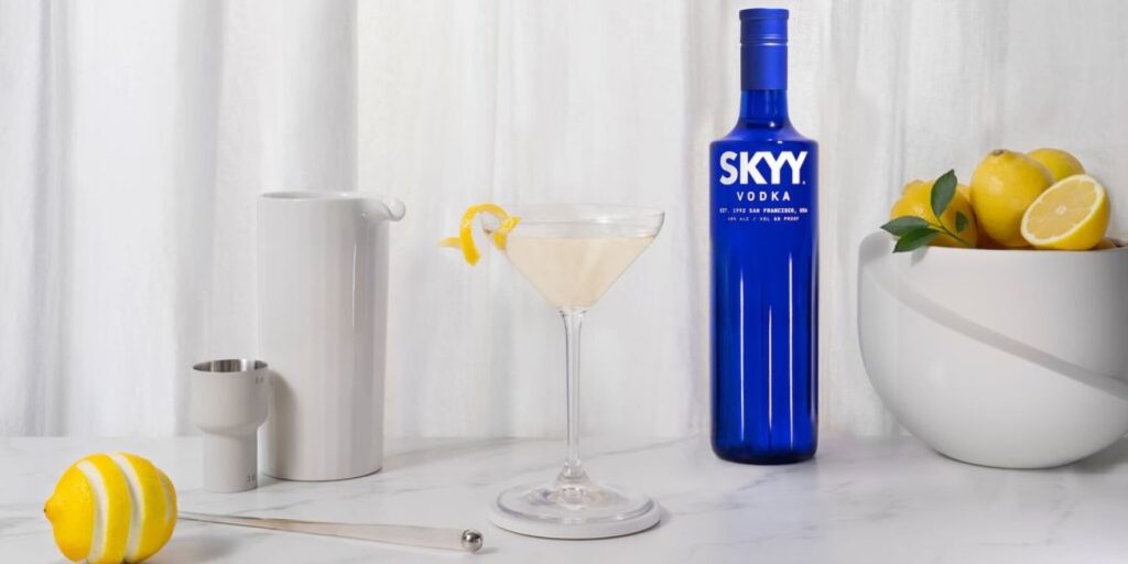 A bottle of SKYY Vodka stands next to a classic vodka martini garnished with a lemon twist. The elegant white backdrop and neatly arranged bar tools give the scene a sophisticated, minimalist appeal. 