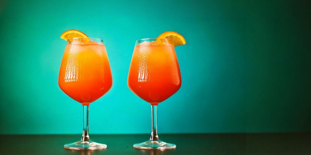 Two glasses of a bright orange cocktail, garnished with slices of orange, sit on a dark surface. The glasses are filled with ice and the drink has a smooth, slushy texture. The background is a gradient of green, creating a striking contrast with the orange drink.