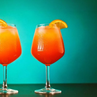 Two glasses of a bright orange cocktail, garnished with slices of orange, sit on a dark surface. The glasses are filled with ice and the drink has a smooth, slushy texture. The background is a gradient of green, creating a striking contrast with the orange drink.