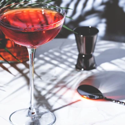 A vibrant sherry cocktail in a coupe glass with red hues, placed on a bright white table with tropical shadows and bar tools in the background.