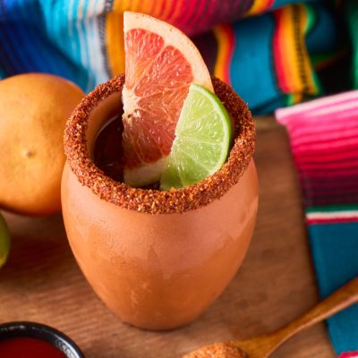 Cantaritos cocktail served in a traditional clay cup with a tajín rim, garnished with grapefruit and lime slices, surrounded by citrus fruit and colourful Mexican textiles, photo by Fabián Montaño/Adobe Stock.