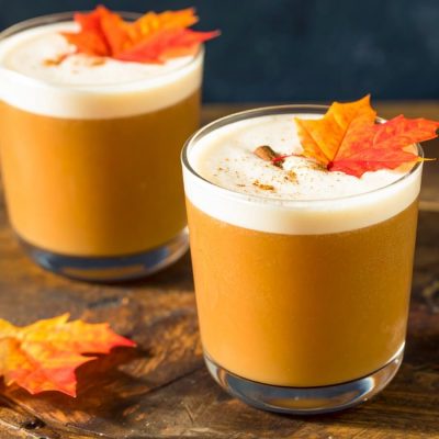 Two autumn-themed maple syrup cocktails with frothy tops in short glasses, garnished with cinnamon and maple leaves, set on a wooden surface.