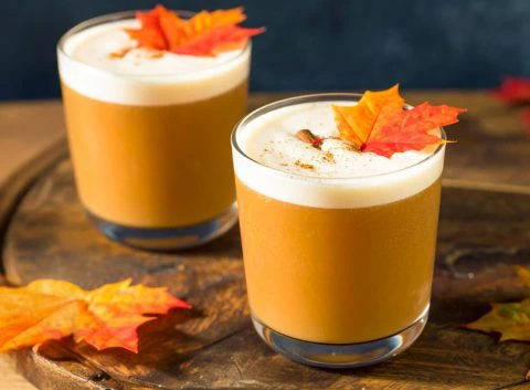 Two autumn-themed maple syrup cocktails with frothy tops in short glasses, garnished with cinnamon and maple leaves, set on a wooden surface.