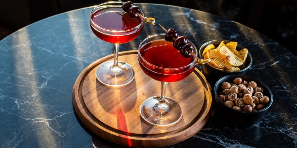 Two Manhattan cocktails with cherry garnishes on a wooden tray set on a marble table