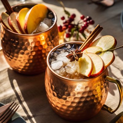 Copper mugs filled with a Thanksgiving batch cocktail of Apple and Cinnamon Mule, garnished with ice, fresh apple slices, cinnamon sticks, and star anise, set on a harvest-themed table with golden napkins and warm lighting.