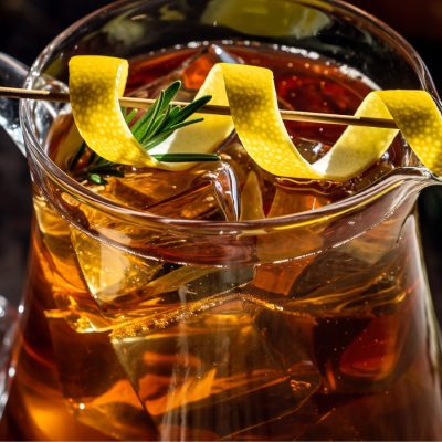 A close-up of a glass pitcher containing Maple Whiskey Sour Punch, a golden-brown Thanksgiving batch cocktail with large ice cubes and garnished with a dramatic lemon peel twist and a sprig of rosemary.