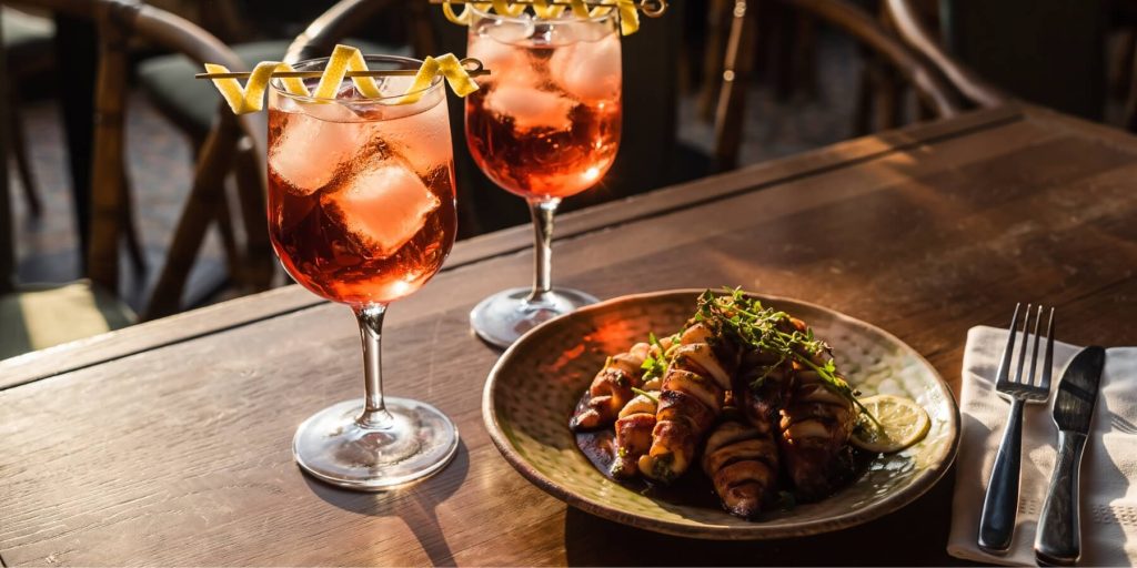 Two pink-hued Vincha Rosada cocktails in stemmed glasses garnished with curled lemon peels, set on a dark wooden table alongside a plate of bacon-wrapped appetizers. The golden-hour light enhances the warm tones of this Argentinian cocktail moment.