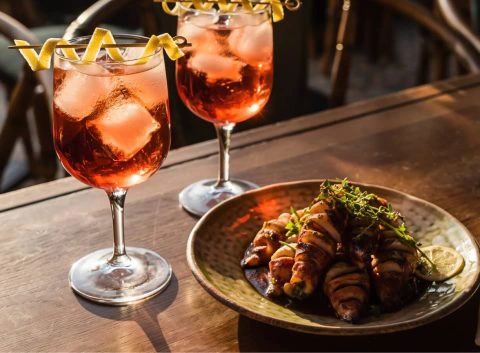 Two pink-hued Vincha Rosada cocktails in stemmed glasses garnished with curled lemon peels, set on a dark wooden table alongside a plate of bacon-wrapped appetizers. The golden-hour light enhances the warm tones of this Argentinian cocktail moment.