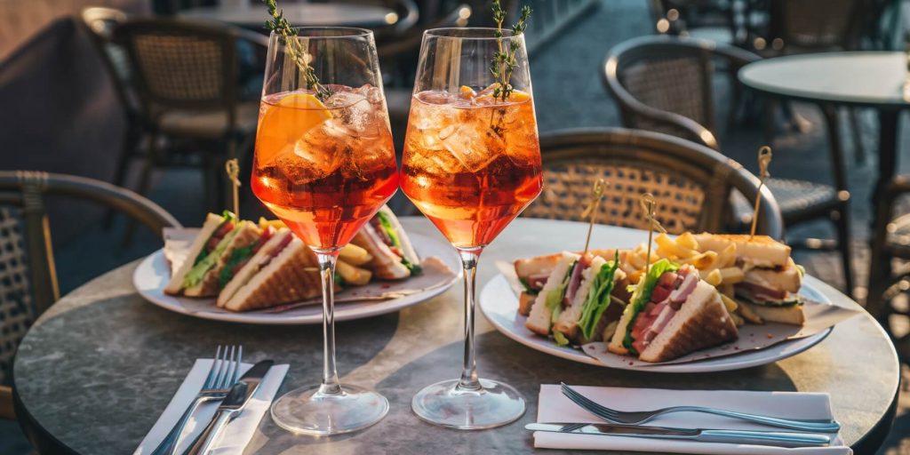 Two spritz cocktails ganished with fresh thyme on a restaurant table with club sandwiches in the background