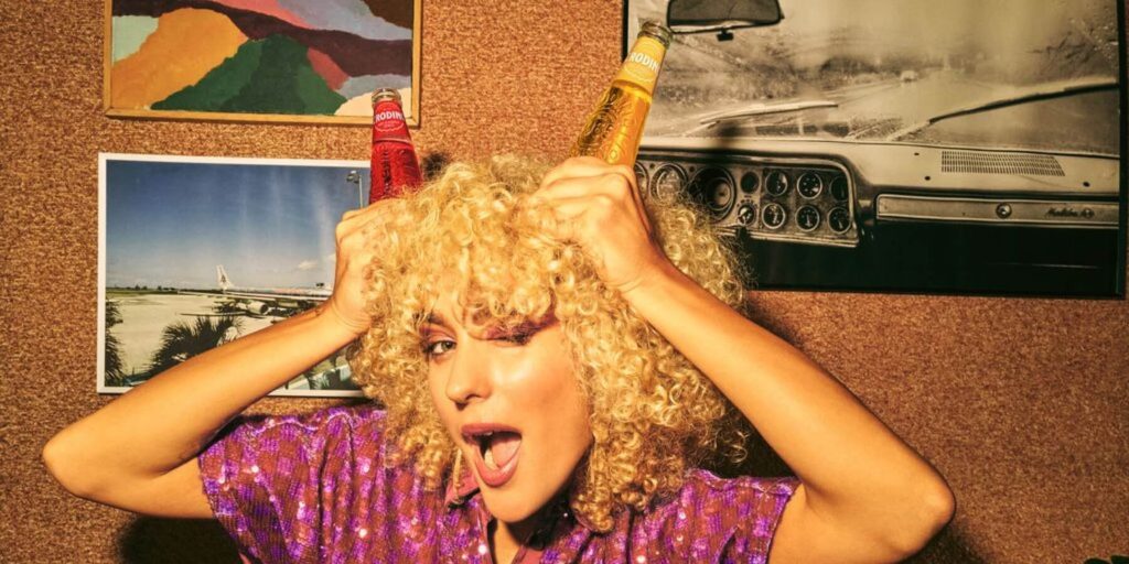 A young lady with blonde curly hair posing with two non-alcholic Crodino bottles on her head