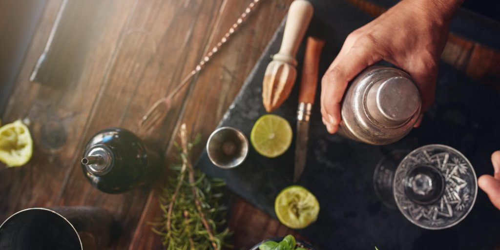 A top shot of a home bar setup with tools and ingredients and a hand holding a cocktail shaker
