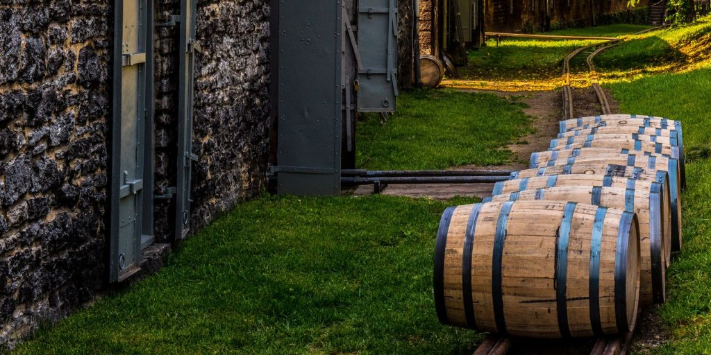 A row of bourbon barrels resting on a track in front of an old stone rickhouse.
