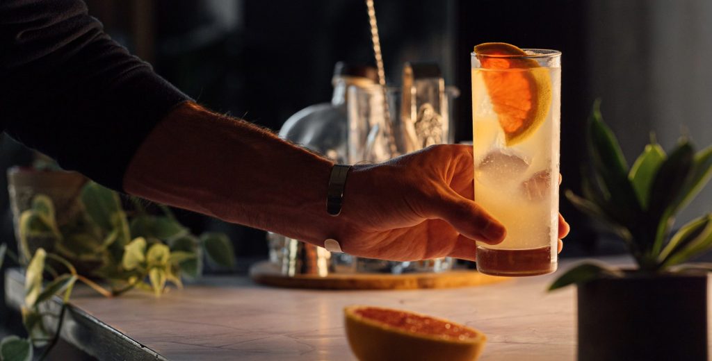 A close up shot of a bartender holding a Paloma cocktail with a home bar set up in the background
