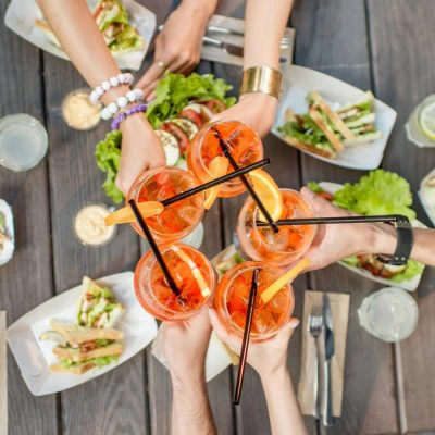 A top shot of a picnic table with salads and sandwiches and a group of friends toasting with Aperol Spritz cocktails.