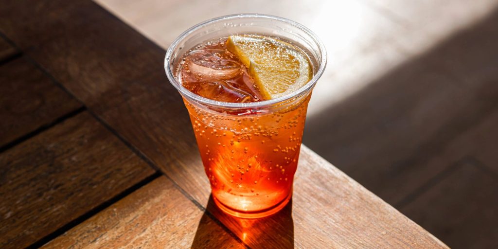 An Aperol Spritz cocktail in a plastic cup on a wooden picnic table.