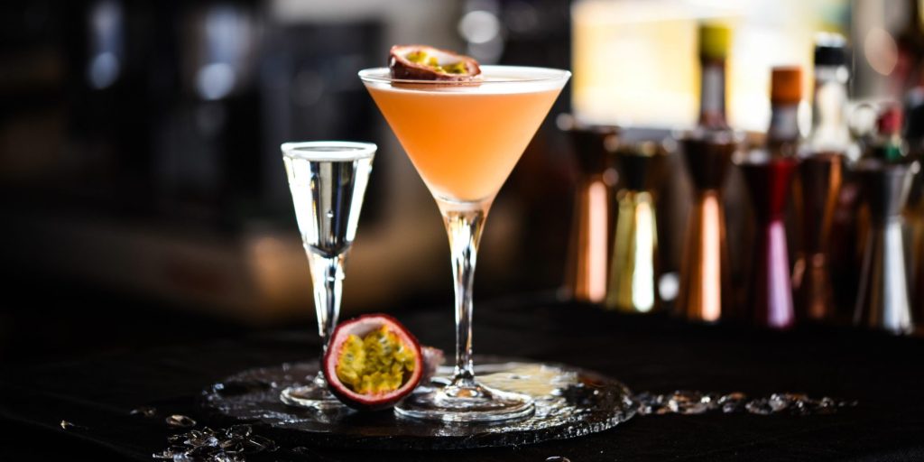 A Pornstar Martini cocktail on a dark bar counter with a half passion fruit next to it and bottles in the background.