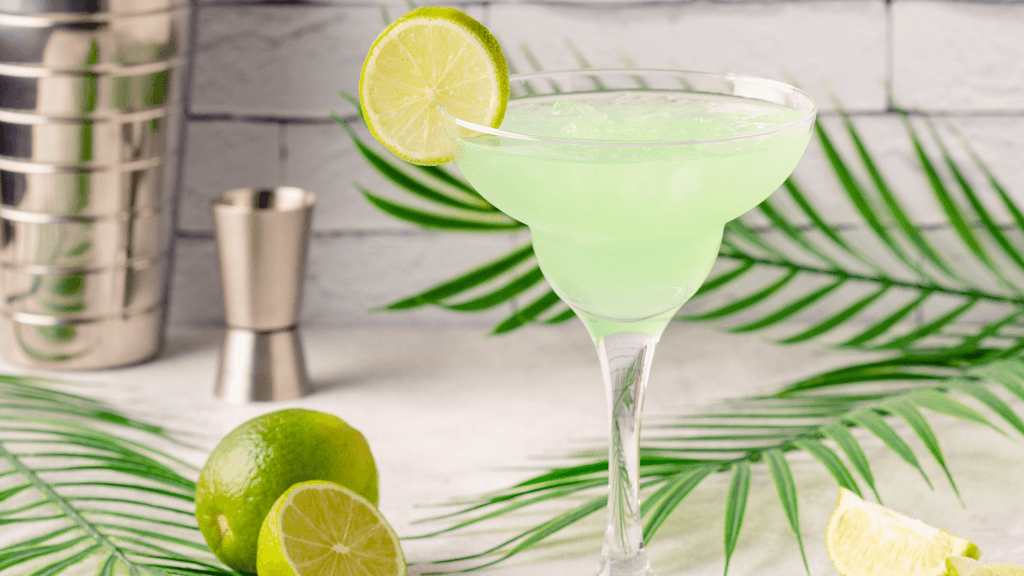 Close up of a Mexican Martini cocktail garnished with a lime wheel, presented against a white backdrop with greenery and fresh limes around it