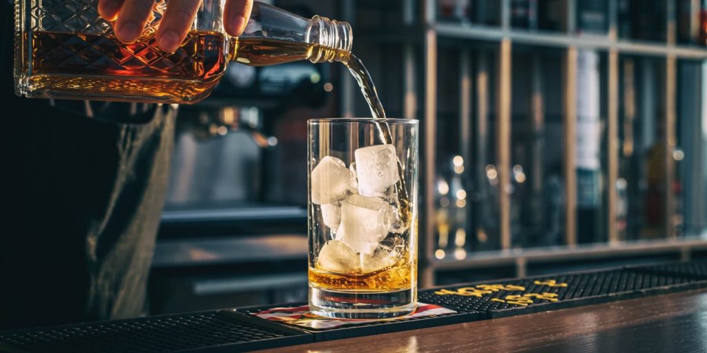 Bartender in a modern bar pouring rum out of a bottle into a tumbler with ice cubes set on a coaster on the bar counter