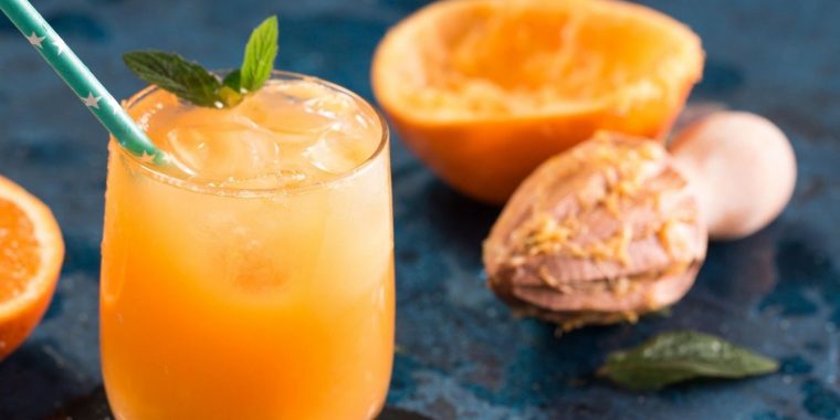 Top view of an Orange Blossom cocktail in a tumbler, garnished with a sprig of mint, featured on a blue surface with an orange in the background