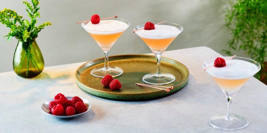 Two frothy pink French Martinis garnished with fresh raspberries placed on a silver serving platter on a table covered in a white tablecloth with a bowl of fresh raspberries and one more French Martini in the foreground