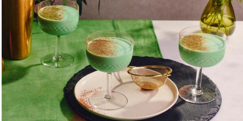 Creamy minty green Grasshopper Cocktails presented on a table covered in a green table cloth, party posed on a white serving platter on top of a slate grey placemat