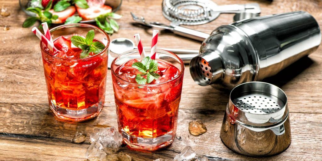 Two bright red mocktails in tumblers with white and red straws with a background of bar tools