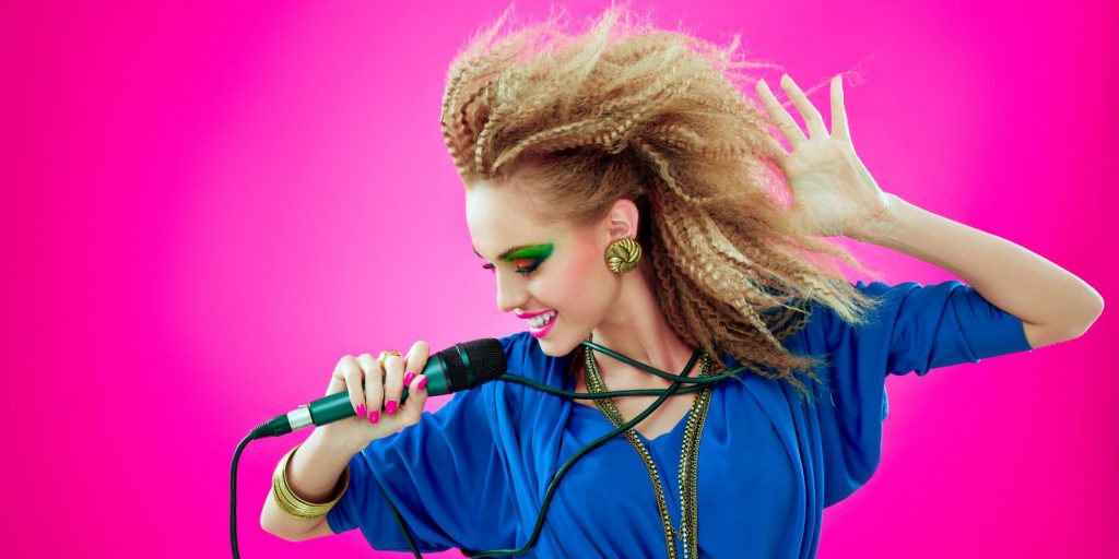 Girl in 80s dress-up clothes singing karoake with crimped hair and birght blue eye shadow against a hot pink backdrop