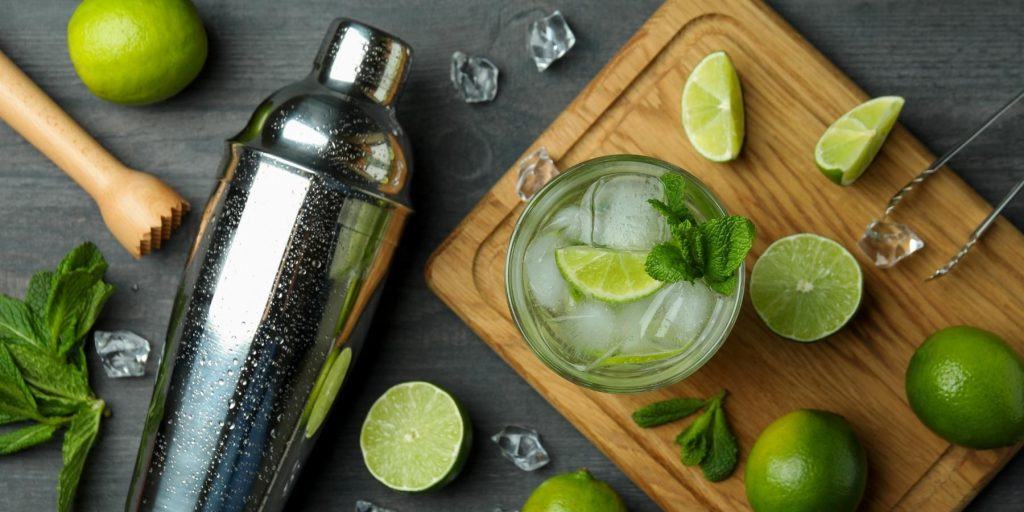 Top shot of lime cocktail on a wooden cutting board, surrounded by fresh lime wedges, flaked by a cocktail shaker, muddler and other cocktail-making tools