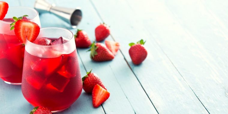 Close up of two Strawberry shrub cocktails in tumbler gasses, garnished with fresh strawberries, presented on a rustic, light blue surface