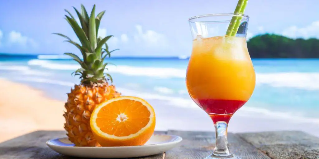 Close-up of a Malibu Sunset cocktail in a Hurricane glass with a view of the ocean in the background and a plate with a sliced orange and fresh pineapple to the left