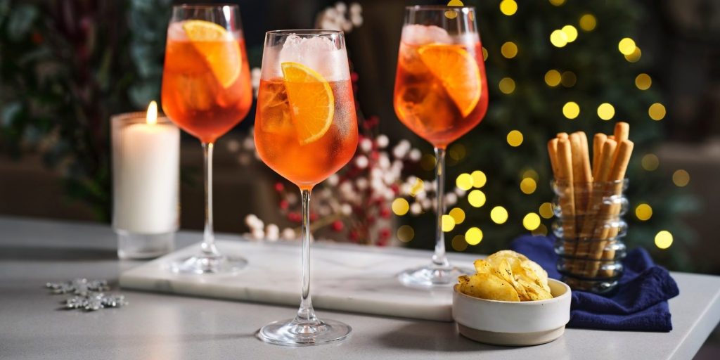 Three Aperol Spritz cocktails in wine glasses garnished with orange slices, viewed from the front, with Christmas trimmings in the background.