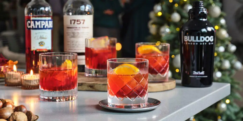 Four Negroni cocktails presented in cut-glass tumblers, placed on a white table among festive Christmas trimmings with a tree in background