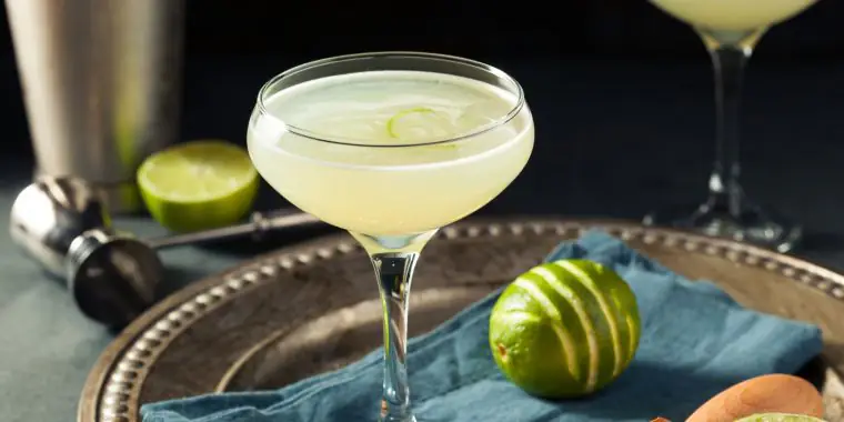 A transluscent Pegu Club cocktail on a pewter serving platter along with a scored lime, against a dark backdrop