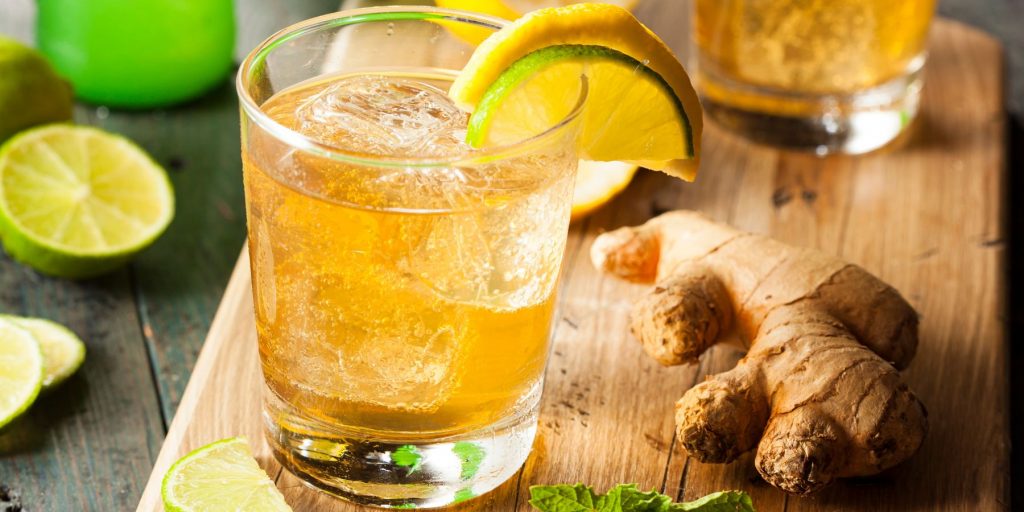 Front view of a Horsefeather Cocktail garnished with slice each of lemon and lime, served on a wooden board with a piece of fresh ginger to the right and sliced limes to the left