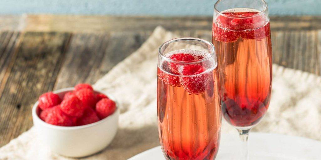 A close-up of two Kir Royale cocktails in Champagne flutes, garnished with fresh raspberries, presented on a wooden surface covered with a white cloth