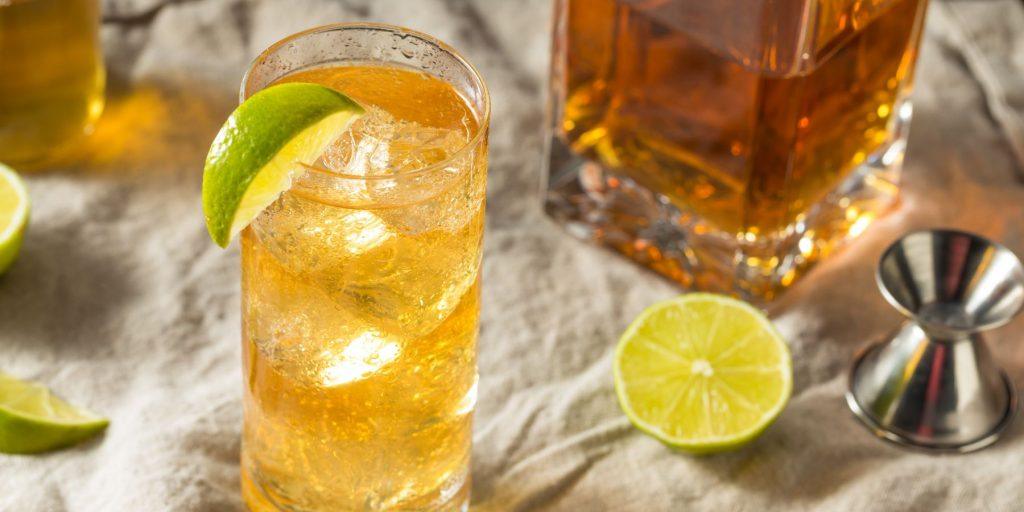 Close-up top shot of a Bourbon Rickey garnished with a piece of lime, with a decanter of bourbon and a sliced lime visible the side