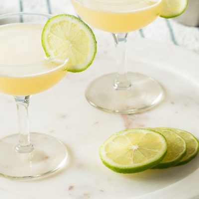 Close up top view of two Gimlet Cocktails in coupe glasses, garnished with lime wheels, and presented on a white platter set on a striped blue and white tablecloth