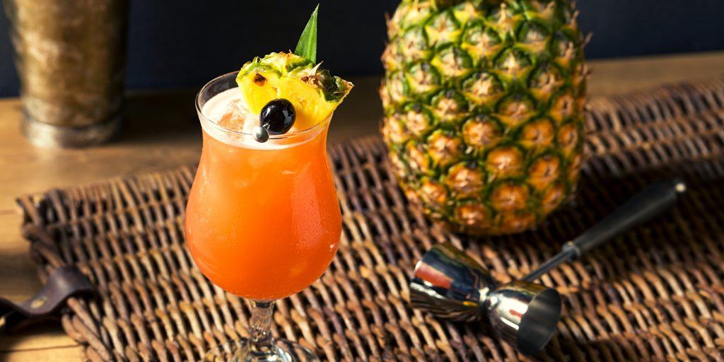 Singapore Sling cocktail with luxardo cherry and pineapple garnish on a woven reed mat on a wooden surface