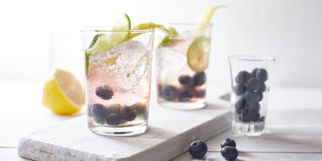 Two Blueberry Lime Spritz cocktails on a white marble countertop against a white backdrop in a light bright home kitchen