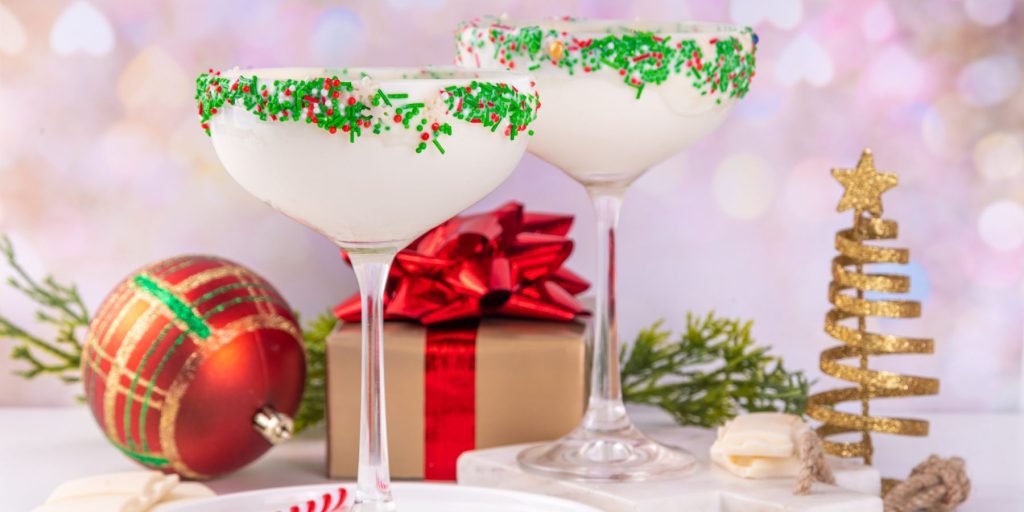 Two Sugar Cookie Martini cocktails with sprinkle rims on a table in the middle of a clutch of decorations and presents for the holidays
